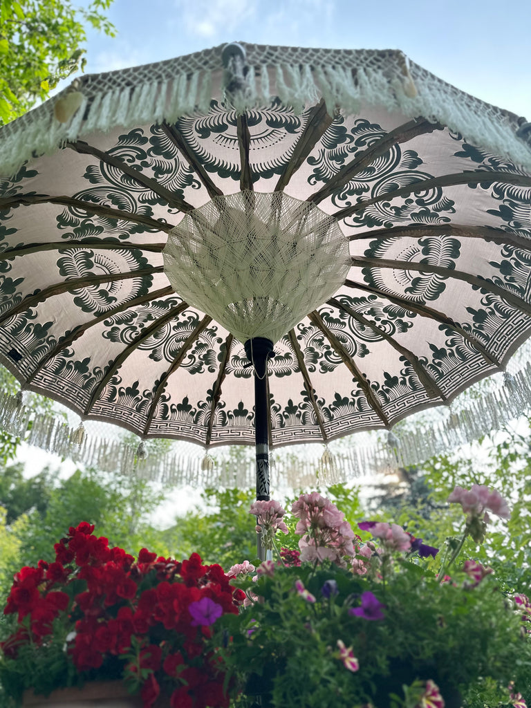 A Baliaric Parasol's Sandy Parasol, featuring intricate patterns and fringed edges, offers shade over vibrant red and pink flowers in a garden setting. Under a clear sky, sunlight filters through the parasol's design, capturing a sense of tropical sophistication.