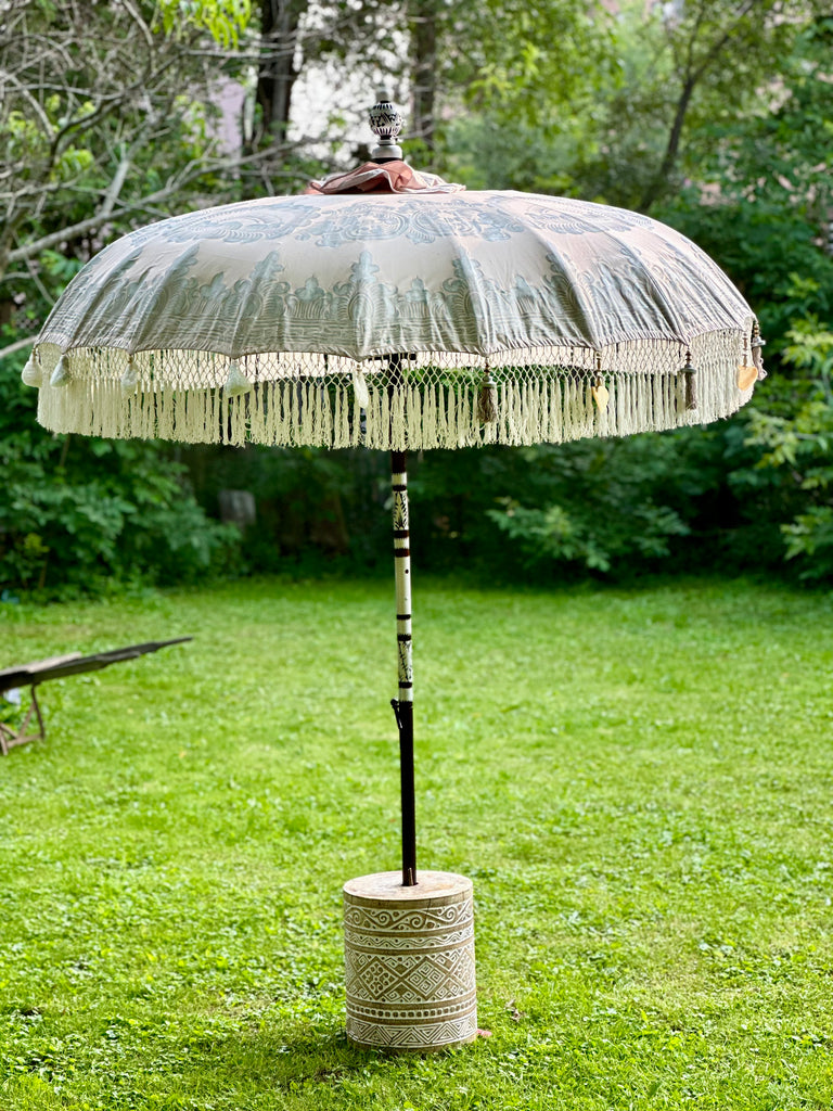 In a grassy garden, the Sandy Parasol by Baliaric Parasol exudes tropical sophistication with its large decorative design complete with tassels. The parasol features a patterned beige canopy and is supported by an ornate pole resting in a carved cylindrical base, all surrounded by lush green foliage.