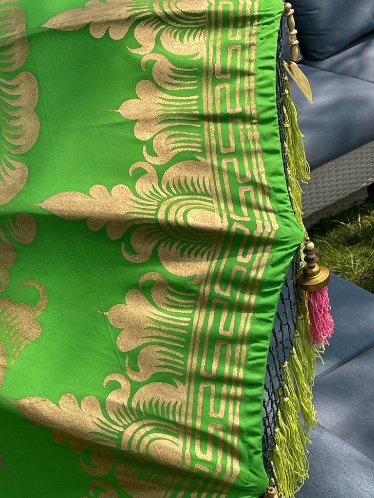 A close-up of the vibrant Samantha Parasol by Baliaric Parasol showcases its intricate gold patterns and designs that exude tropical elegance. Pink and yellow tassels hang from the edge, with a glimpse of a cushioned gray seat in the background adding to its handcrafted charm.