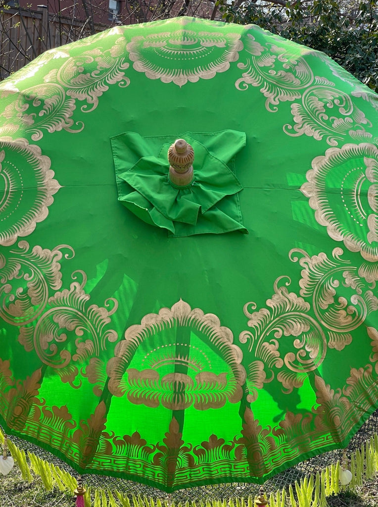 The Samantha Parasol from Baliaric Parasol is a vibrant green umbrella adorned with intricate gold patterns featuring floral designs and tassels along the edges. When viewed from above, this handcrafted parasol opens to reveal its symmetrical elegance and exquisite detail, capturing tropical sophistication in every motif.