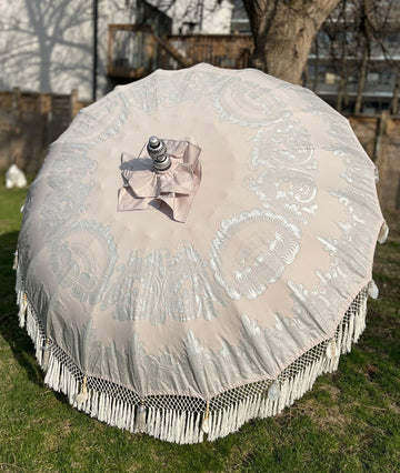 A large, beige outdoor umbrella from Baliaric Parasol, named the Sandy Parasol, stands open on a grassy lawn. Its charm is enhanced by intricate white lace patterns and fringe edging. Nearby are trees and a wooden fence, with a house visible in the background. Shadows cast on the grass amplify its tropical sophistication.