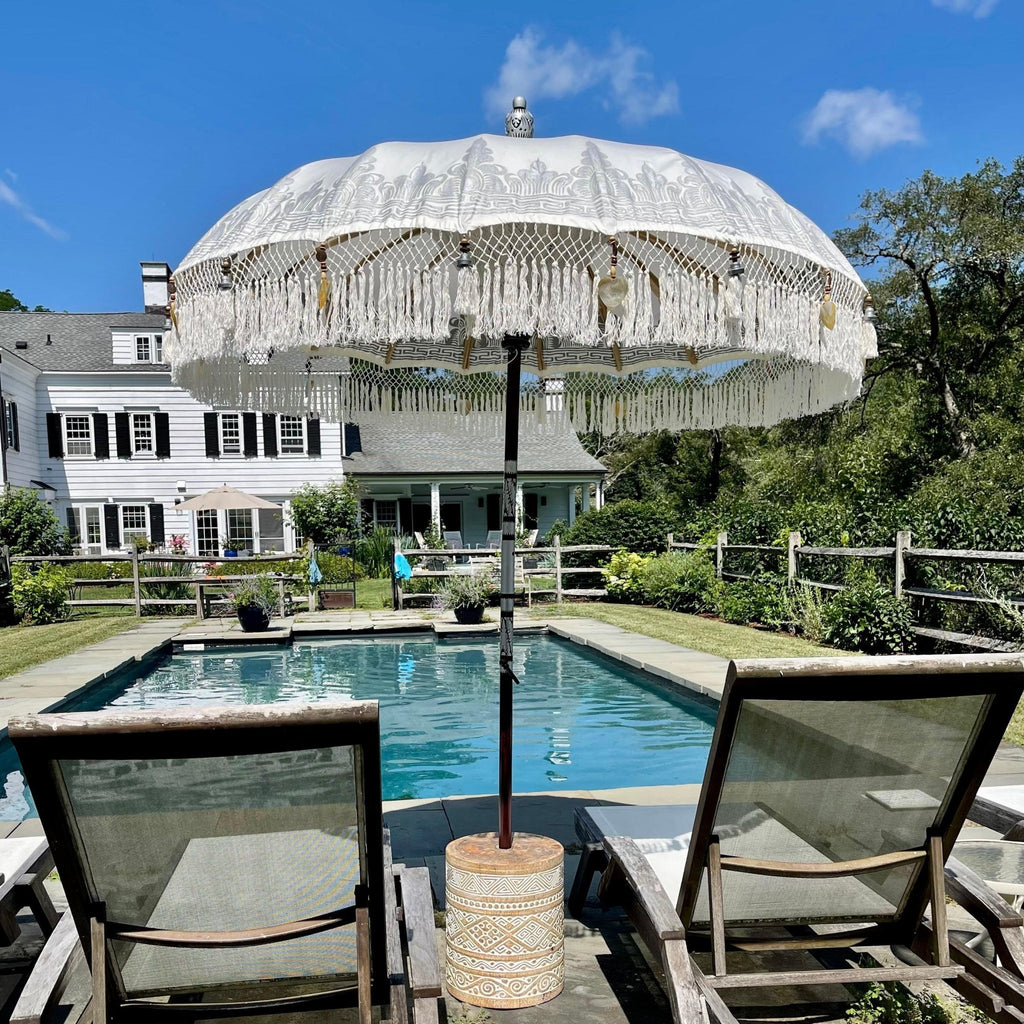 A scenic backyard features a swimming pool surrounded by greenery and a white, two-story house. Two lounge chairs with a decorative White Cloud Parasol from Baliaric Parasol grace the foreground, offering shade and a relaxing view of the calm pool and clear blue sky.