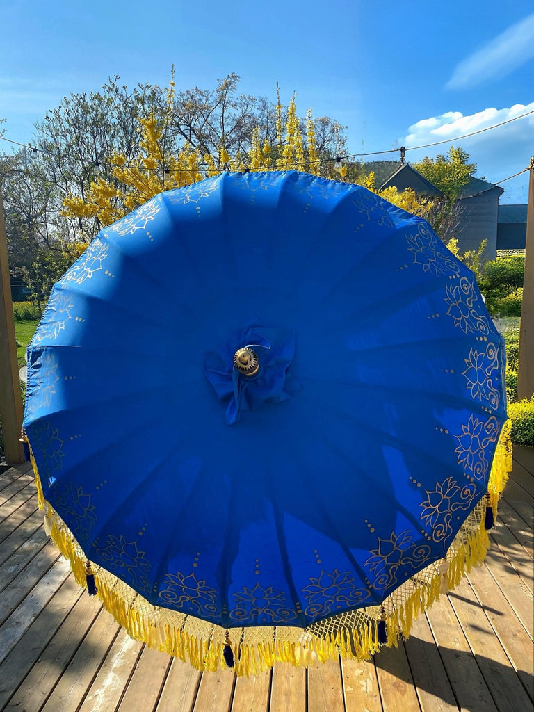 A large, ornate blue Carina Parasol by Baliaric Parasol, adorned with golden fringes and floral patterns, is open on a wooden deck, contributing an element of elegance to the outdoor decor. In the background are trees and a clear blue sky with a few clouds.
